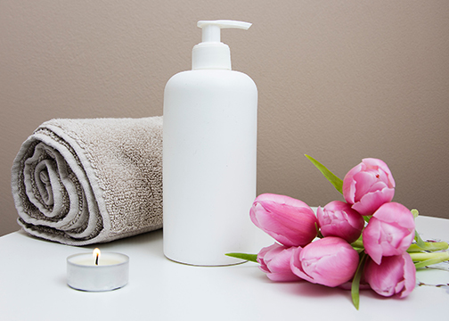 white bottle of lotion next to tan towl, a lit tealight candle, and bright pink tulips