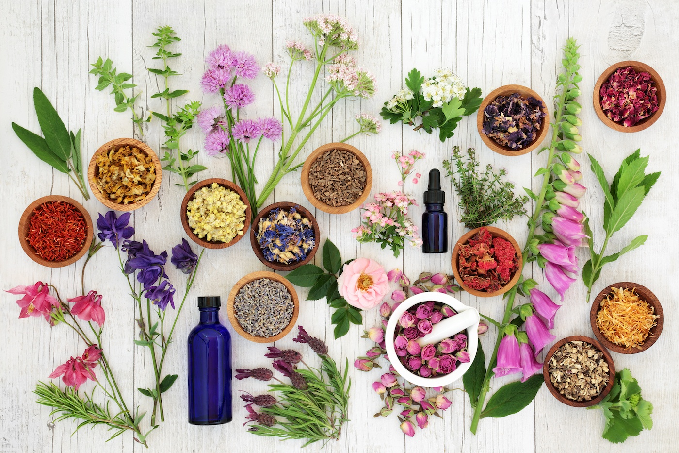 essential oil bottle and various flowers and herbs laying on a white wood table